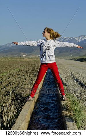 Stock Photograph - Teenage girl standing over a small canal of water with arms outstretched; Andalusia, Spain. Fotosearch