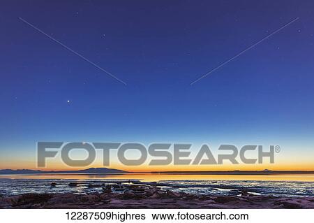 Stock Photograph - The moon rises above Mount Susitna on a clear winter night, sea ice visible in the foreground; Anchorage, Alaska, United States of America. Fotosearch