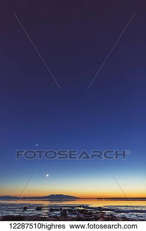 Stock Photograph - The moon rises above Mount Susitna on a clear winter night, sea ice visible in the foreground; Anchorage, Alaska, United States of America. Fotosearch