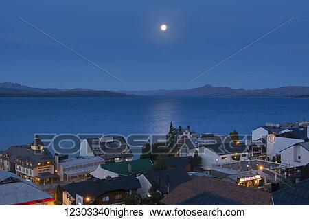 Stock Photograph - The moon rising over a lake from above the town roof tops showing some yellow city lights against the blue scene; Bariloche, Argentina. Fotosearch