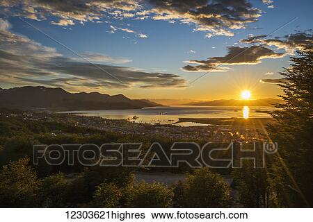 Stock Photograph - The sun rises over the port of Ushuaia in Argentina as a ship enters the calm bay, trees in the foreground and low hills on the horizon, while the sky is dotted with clouds and turns from a golden yellow to a deep blue; Ushuaia, Argentina. Fotosearch