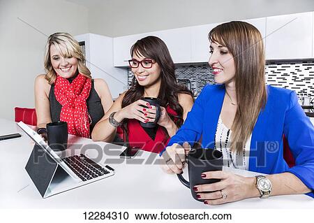 Stock Image - Three professional business women looking on social media while taking a break in an office kitchen area; St. Albert, Alberta, Canada. Fotosearch
