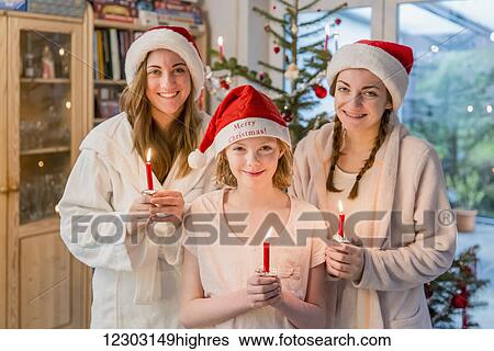 Stock Photograph - Three sisters wearing santa hats and holding lit candles standing in front of a Christmas tree decorated with ornaments and tinsel; Bonn, Nordrhein Westfalen, Germany. Fotosearch