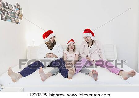 Three sisters wearing santa hats, pajamas and robes and sitting together on a couch looking at two cell phones while smiling; Bonn, Nordrhein Westfalen, Germany View Large Photo Image Stock Photograph - Three sisters wearing santa hats, pajamas and robes and sitting together on a couch looking at two cell phones while smiling; Bonn, Nordrhein Westfalen, Germany. Fotosearch