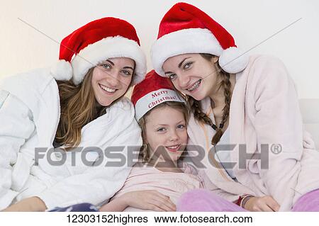 Stock Photograph - Three sisters wearing santa hats, pajamas and robes and sitting together on a couch; Bonn, Nordrhein Westfalen, Germany. Fotosearch