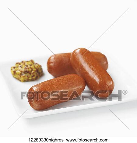 Stock Photograph - Three small wieners on a plate with a grainy mustard; Toronto, Ontario, Canada. Fotosearch