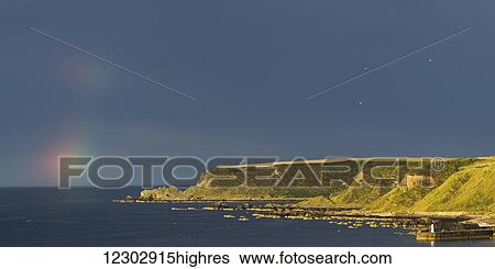 Stock Photograph - Traces of a rainbow in the sky in the distance over the ocean with cliffs along the coastline; Cullen, Moray, Scotland. Fotosearch