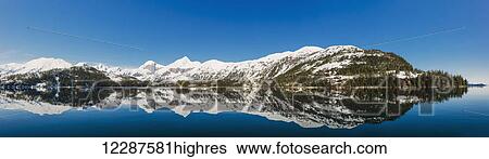 Stock Photograph - Tranquil ocean waters reflect snow covered mountains on a clear day in Kings Bay, Prince William Sound; Whittier, Alaska, United States of America. Fotosearch