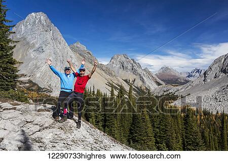Stock Photograph - Two male hikers with arms raised in the air on rock ridge overlooking alpine valley with colourful larch trees in autumn and rocky peaks with blue sky and clouds, Kananaskis Provincial Park; Alberta, Canada. Fotosearch