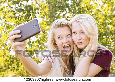 Stock Photograph - Two sisters having fun outdoors in a city park in autumn and taking selfies of themselves and making funny faces; Edmonton, Alberta, Canada. Fotosearch