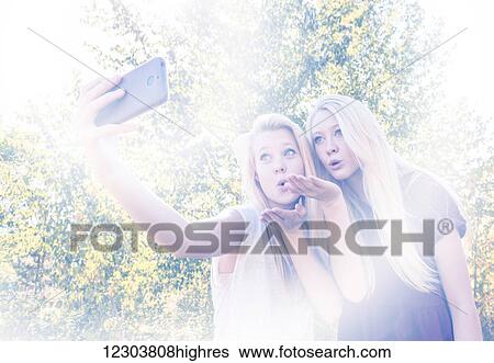 Stock Photograph - Two sisters having fun outdoors in a city park in autumn and taking selfies of themselves and blowing kisses; Edmonton, Alberta, Canada. Fotosearch