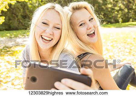 Stock Photograph - Two sisters having fun outdoors in a city park in autumn and laughing at the selfies they have taken of themselves; Edmonton, Alberta, Canada. Fotosearch