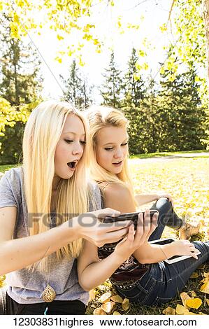 Stock Photograph - Two sisters having fun outdoors in a city park and looking at social media on their smart phone and tablet; Edmonton, Alberta, Canada. Fotosearch