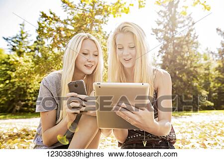 Two sisters having fun outdoors in a city park in autumn looking at social media on their smart phone and tablet; Edmonton, Alberta, Canada View Large Photo Image Stock Photograph - Two sisters having fun outdoors in a city park in autumn looking at social media on their smart phone and tablet; Edmonton, Alberta, Canada. Fotosearch