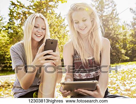 Stock Photograph - Two sisters in a city park enjoying time together looking at social media on their smart phone and tablet; Edmonton, Alberta, Canada. Fotosearch