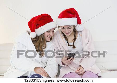 Stock Photograph - Two teenage sisters wearing santa hats, pajamas and robes and sitting together on a couch looking at a cell phone; Bonn, Nordrhein Westfalen, Germany. Fotosearch