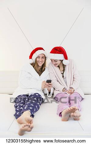 Stock Photograph - Two teenage sisters wearing santa hats, pajamas and robes and sitting together on a couch looking at a cell phone; Bonn, Nordrhein Westfalen, Germany. Fotosearch