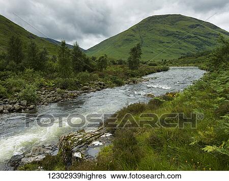 Stock Photograph - Water flowing through lush green hills under a cloudy sky; Scotland. Fotosearch
