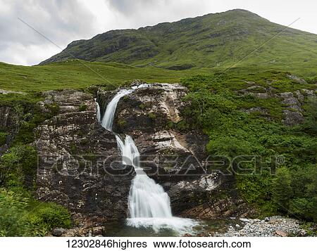 Stock Photograph - Waterfall over a rugged rock cliff with lush green grass on the landscape under a cloudy sky; Scotland. Fotosearch