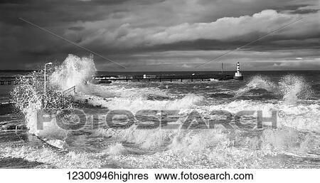 Stock Photograph - Waves crashing along the coast with a small lighthouse at the end of a pier; Amble, Northumberland, England. Fotosearch