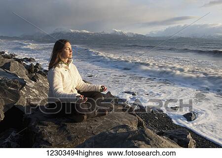 Stock Photograph - Woman practicing meditation on a rocky beach, Homer Spit, Southcentral Alaska. Fotosearch