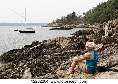 Woman sitting on rocks at the water's edge looking out over the coast; Bowen Island, British Columbia, Canada View Large Photo Image Picture - Woman sitting on rocks at the water's edge looking out over the coast; Bowen Island, British Columbia, Canada. Fotosearch