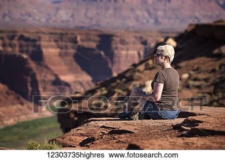 Stock Photograph - Woman sitting on the edge of a rock canyon cliff looking over the Colorado river with canyon walls in the background; Colorado, United States of America. Fotosearch