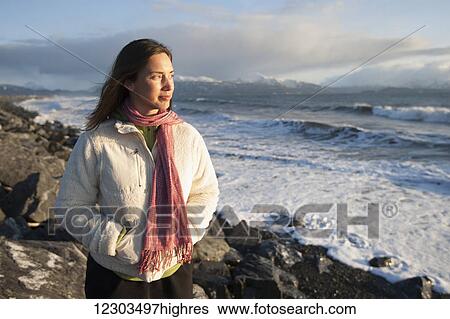 Stock Photograph - Woman standing along the rocky shoreline of Homer Spit at sunset in winter, Southcentral Alaska. Fotosearch