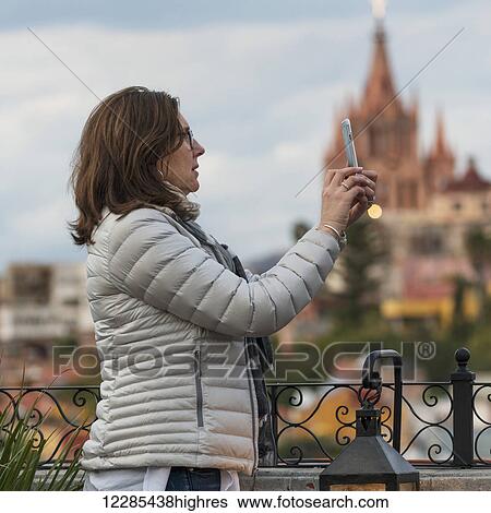 Stock Photograph - Woman taking a photograph with a smart phone and a church in the distance; San Miguel de Allende, Guanajuato, Mexico. Fotosearch
