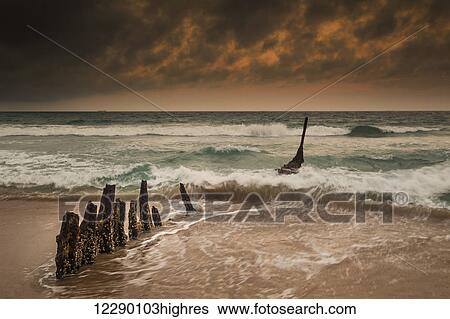 Wooden posts on a beach with a boat being tossed in the water and waves crashing into the sand under a cloudy sky; Queensland, Australia View Large Photo Image Stock Photograph - Wooden posts on a beach with a boat being tossed in the water and waves crashing into the sand under a cloudy sky; Queensland, Australia. Fotosearch