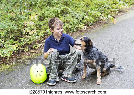 Stock Photograph - Young boy sitting on a skateboard and petting his dog; Salmon Arm, British Columbia, Canada. Fotosearch
