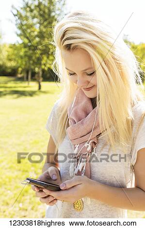 Stock Photograph - Young woman with long hair listening to music outdoors on her smart phone in a city park in autumn; Edmonton, Alberta, Canada. Fotosearch
