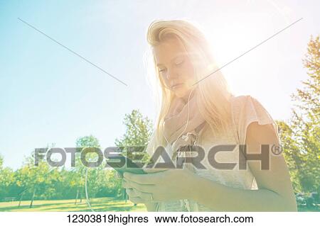Stock Photograph - Young woman with long hair listening to music outdoors on her smart phone in a city park in autumn; Edmonton, Alberta, Canada. Fotosearch