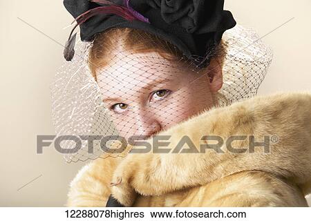 Stock Photograph - Young woman with red hair in a veiled black hat with feathers and fur coat, mouth hidden; Caldecott, England. Fotosearch