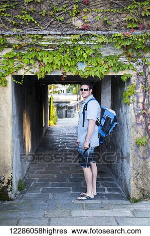 Stock Photograph - A backpacking young male tourist walks beneath a historic village tunnel in Lake Como in summer attire; Varenna, Lombardy, Italy. Fotosearch