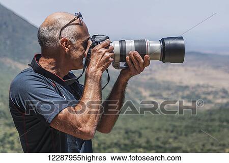 Stock Photograph - A balding man is photographing a view of the Rift Valley in the sunshine using a digital SLR camera with a long lens; Tharaka Nithi, Kenya. Fotosearch