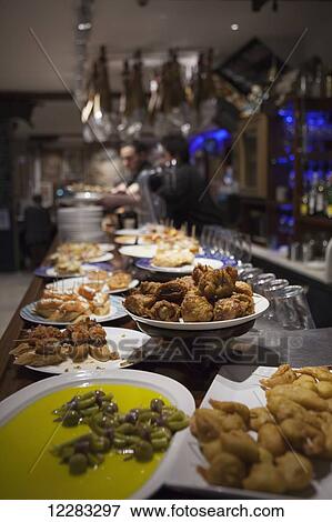 A bar man stands underneath hanging legs of ham (Jamon iberico) with a spread of pintxos on the bar counter; San Sebastian, Spain View Large Photo Image Stock Photo - A bar man stands underneath hanging legs of ham (Jamon iberico) with a spread of pintxos on the bar counter; San Sebastian, Spain. Fotosearch