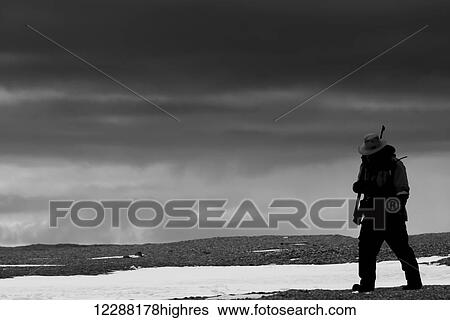 Stock Photograph - A black and white image of a man walking over a rocky Arctic snowfield with a rifle over his shoulder; Spitsbergen, Svalbard, Norway. Fotosearch