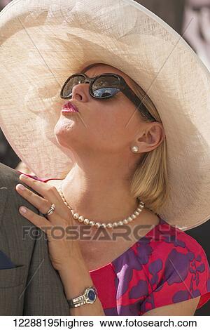 Stock Photograph - A blonde woman in a colourful red and purple dress, sunglasses and a mesh hat is pouting at her husband; Ascot, England. Fotosearch