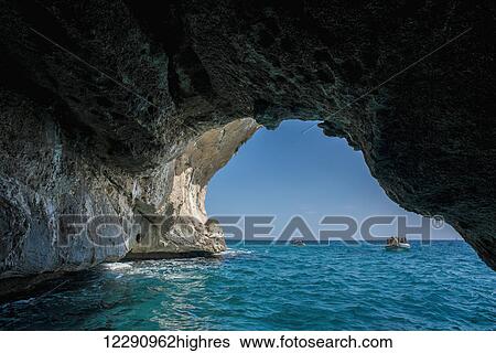 Stock Photograph - A boat of tourists approaching a cave along the coast; Cala Gonone, Sardinia, Italy. Fotosearch