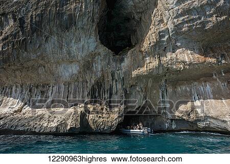 Stock Photograph - A boat of tourists approaching a cave along the coast; Cala Gonone, Sardinia, Italy. Fotosearch