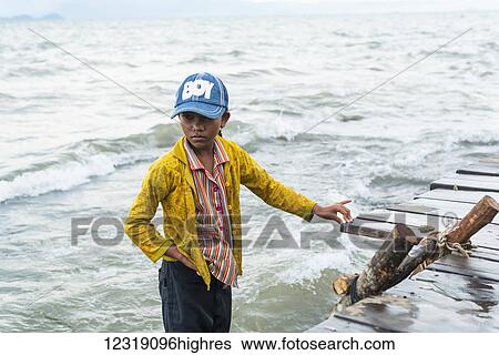Stock Photograph - A boy stands in the water at the edge of the dock, local life in the famous crab market; Kep, Cambodia. Fotosearch
