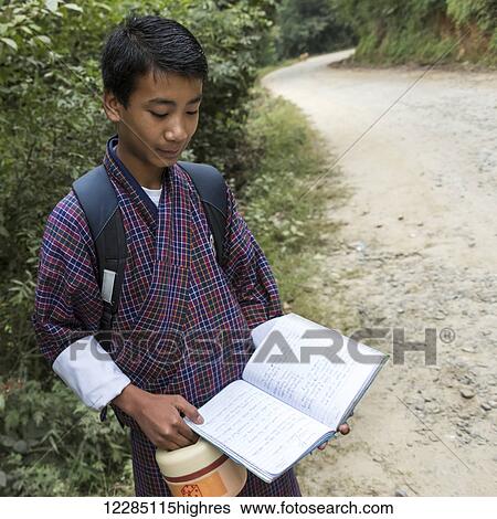 Stock Photograph - A boy with a checkered robe carrying a backpack and reading a book on the side of a road; Punakha, Bhutan. Fotosearch