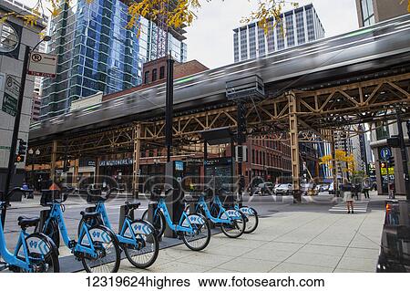 Stock Photograph - A Chicago Transit Authority L train traversing The Loop passes the DIVVY bikeshare station at Franklin and Lake Streets; Chicago, Illi0is, United States of America. Fotosearch