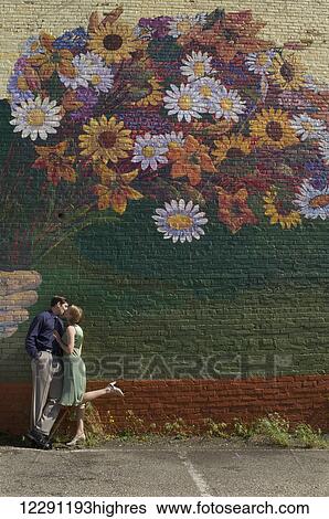 Stock Photograph - A couple kisses as they lean against a wall with a large mural of a colourful bouquet of flowers painted on the wall; Raleigh, North Carolina, United States of America. Fotosearch