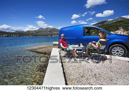 Stock Photograph - A couple sits outside their camper van at a beach along the coast; Slano, Croatia. Fotosearch
