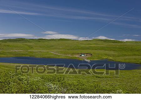 Stock Photograph - A dirt road leads to a small green warehouse and dock on a small Lake, Sand Point, Popof Island, Southwestern Alaska, USA, Summer. Fotosearch