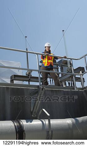 Stock Photograph - A female civil engineer, wearing a hard hat and a reflective orange vest, stands on a walkway in front of a water filtration system; Chapel Hill, North Carolina, United States of America. Fotosearch