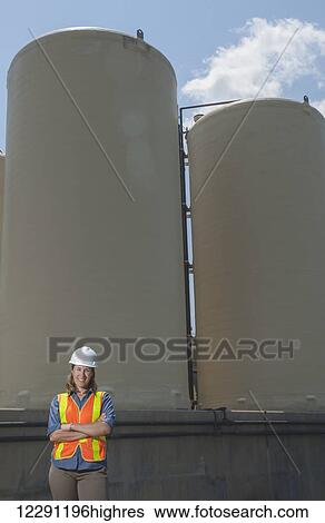 Stock Photograph - A female civil engineer, wearing a hard hat and a reflective orange vest, stands in front of two water filtration tanks; Chapel Hill, North Carolina, Untied States of America. Fotosearch