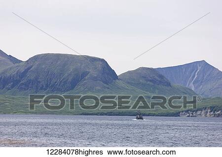 Stock Photograph - A fishing boat motors out of the Sand Point Harbor towards distant mountains, Sand Point, Southwestern Alaska, USA, Summer. Fotosearch